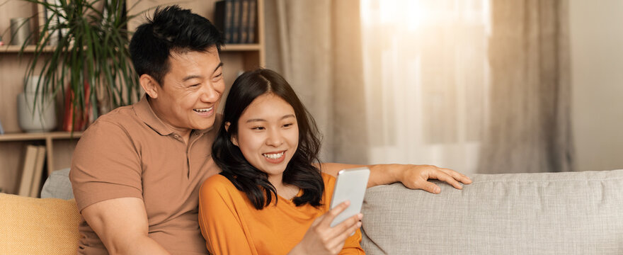 Happy Asian Couple, Young Adults, Sitting Close Together On Couch, Sharing Laughter While Using Smartphone At Home, Enjoying Time Together In Cozy Living Room. Lifestyle Concept.