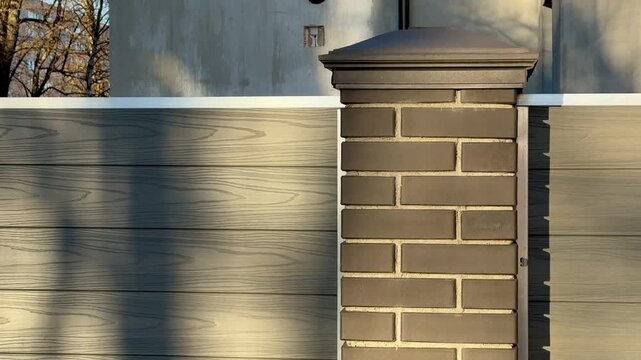 Sunlit brick column by cottage fence, warm golden hour light casts long tree shadows across weathered wooden panels, capstone