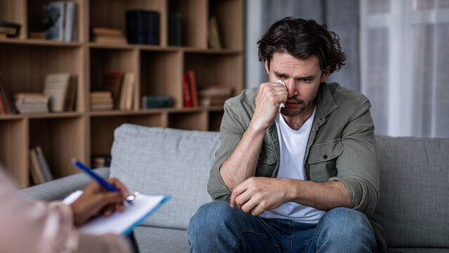 Worried Caucasian Man In His 30s Sitting On Couch During Therapy Session, Discussing Emotions With Therapist In Office Environment, Seeking Guidance, Mental Health Concept.