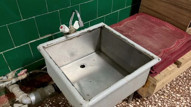 Rusty iron foot sink under faucet, set against green ceramic tiles, exposed pipes and valve, stained basin floor with clogged