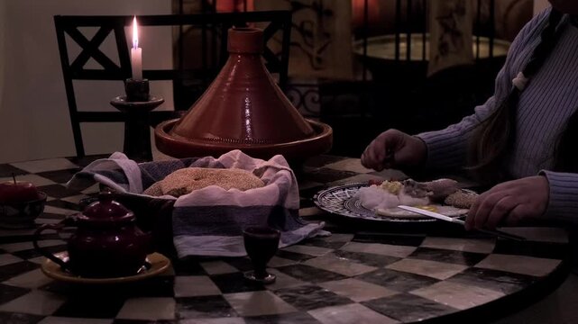 Woman eating at Moroccan table, tagine and bread, cakes, and tea, candle burning, cultural dining experience, Moroccan ceramic plates, representing traditional cuisine and hospitality