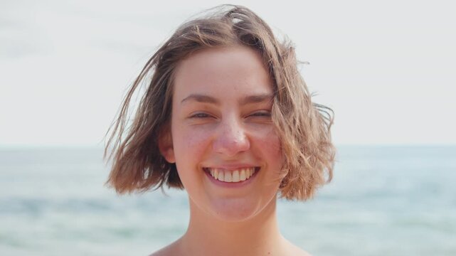 Young woman smiling at camera with wind blowing through her hair enjoying fresh air against sea representing positive energy, happiness and confidence. Closeup portrait