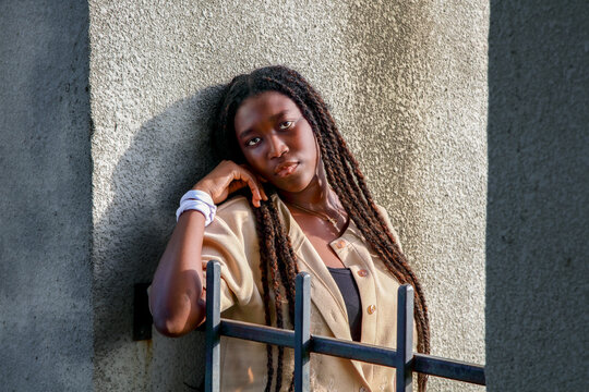 Urban lifestyle portrait of a young Black woman leaning on a concrete wall behind a metal fence in warm sunlight