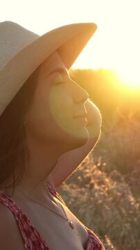 Profile view of a beautiful young woman with a hat smiling and enjoying the warm golden light of the sun during a summer sunset in a countryside field, feeling freedom and happiness