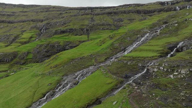 A push in aerial drone shot shows a hiker walking on a remote mountain trail with multiple waterfalls cascading down, showcasing the lush, rugged landscape on the Faroe Islands.