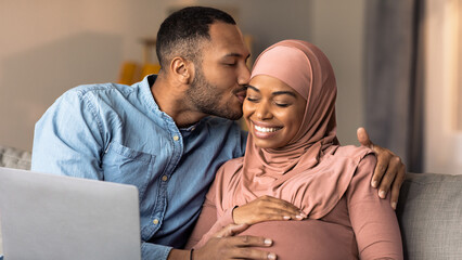 Obraz premium Joyful Embracing Expecting African American Couple Smiling Together Sitting On Sofa At Home, Engaged In Warm Affection, Celebrating Pregnancy Journey With Love And Anticipation. Family Concept.