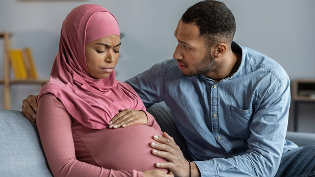 Worried Black Woman With Pink Hijab Holding Pregnant Belly While Partner Comforts Her At Home, Showing Concern. Intimate Moment Between Couple In A Comforting Setting, Pregnancy Concept.