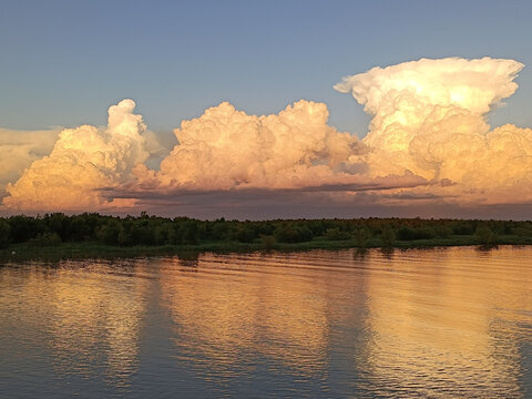 Cloudy landscape at sunset by the river. Nature background with beautiful sky, green trees and bright river.