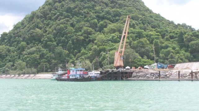 workboat moored beside a dock with a crane along the tropical limestone shoreline of Khanom, Thailand, featuring calm turquoise water and lush green hills