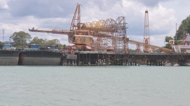 industrial waterfront terminal with conveyor loading equipment, cargo barges, and marine infrastructure along the gulf of thailand at khanom, thailand under a dramatic cloudy sky