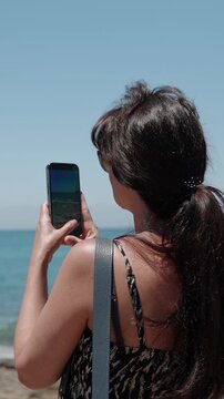 Back view of adult woman filming the sea with smartphone on sunny beach. Travel lifestyle scene with ocean view, mobile video content, summer tourism, social media and vacation memories.
