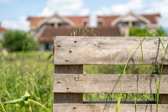 old, dilapidated wooden fence stands in tall green grass, with a rural house and pine tree visible in the blurred background