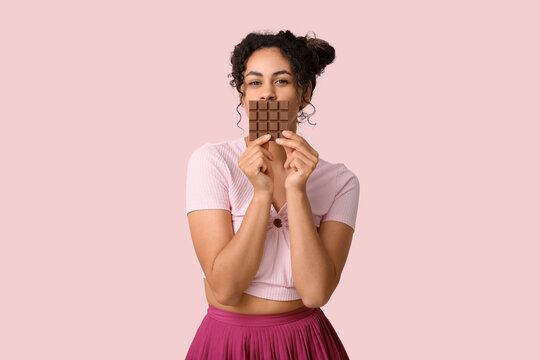 Young African-American woman with bar of chocolate on pink background