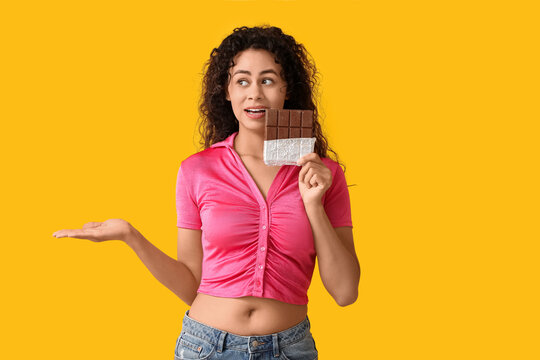 Beautiful young African-American woman with sweet chocolate bar showing something on yellow background