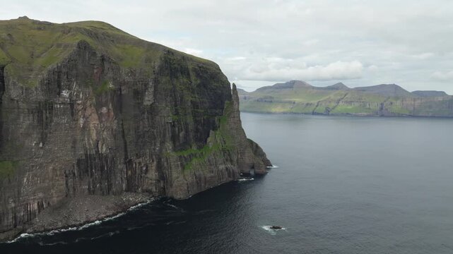A drone shot panning right over rugged cliffs called Tr&oslash;llkonufingur (The Witch&rsquo;s Finger) and green mountains meeting the sea on the Faroe Islands. Capturing the remote, dramatic coastal landscape.