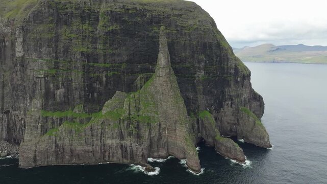 Aerial drone footage panning right, showcasing rugged cliffs called Tr&oslash;llkonufingur (The Witch&rsquo;s Finger) on the Faroe Islands. Overcast skies highlight the dramatic landscape and remote beauty.