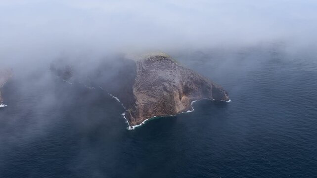 Aerial view of Ilheus das Cabras, rocky islets rising from the Atlantic Ocean, partially obscured by low-hanging clouds and mist in Azores, Portugal.