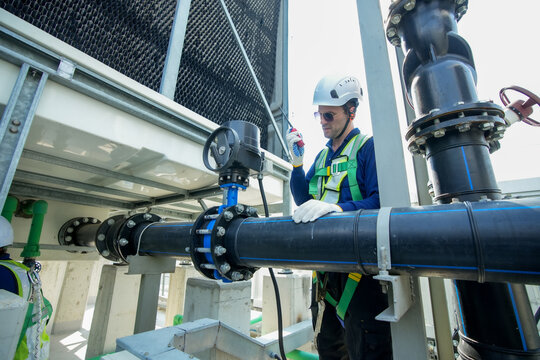 Engineer maintenance hvac cooling tower technician inspection rooftop adjusts valve and gauge on large pipe with safety harness and hard hat for routine service, ensuring reliable water flow