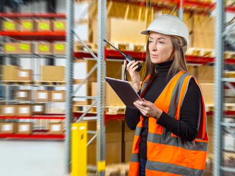 Warehouse logistics supervisor coordinates inventory using walkie-talkie and tablet in distribution center aisle with stocked warehouse shelves