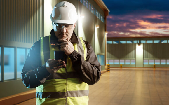 Industrial logistics supervisor reviews operations using smartphone at warehouse facility during evening inspection with safety gear and reflective vest