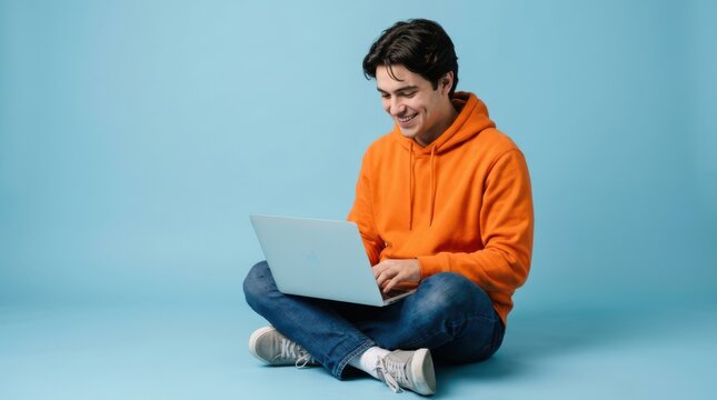 Young man sitting on the floor using a laptop against a blue background