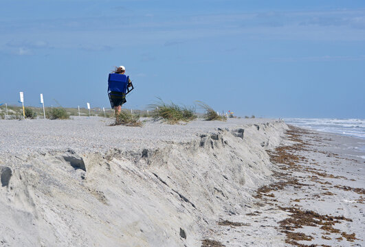 A beachgoer carrying a beach chair backpack strolls walking away along the dunes at St. Augustine Beach, with the shoreline, crashing waves, and the horizon meeting the sky in the distance.