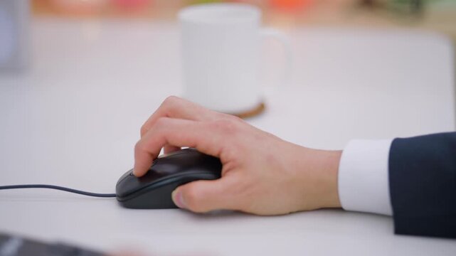 A close-up of a businessman's hand using a computer mouse on a desk in a bright office environment.