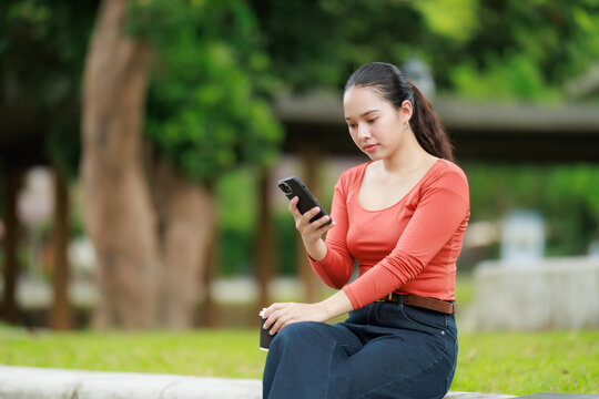 Young Asian woman is sitting outdoors in a park, holding a smartphone and a coffee cup, focusing on her device for communication, social media, or remote work activities