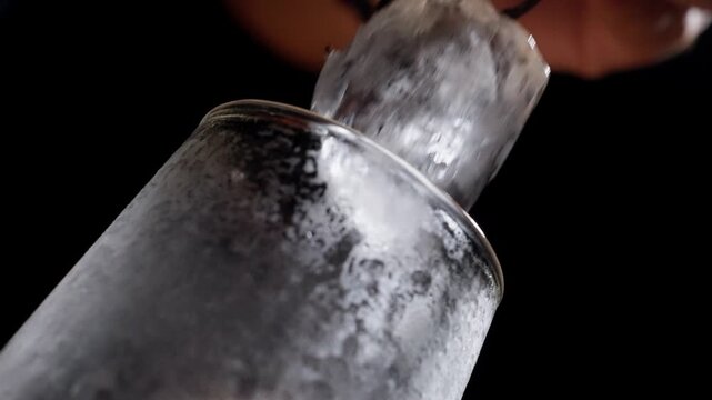 Metal tongs place ice cube into chilled fluted glass against black backdrop