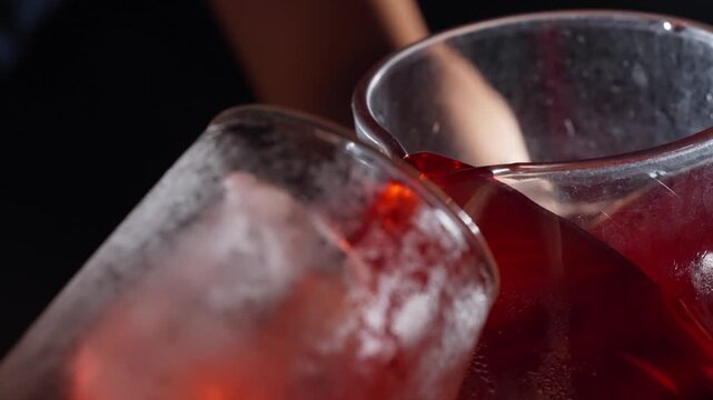 Deep red cocktail pours into chilled glass at angle, smooth slow motion flow jostling ice cube