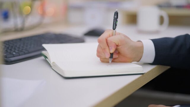 A close-up of a businessman's hand writing on a notepad with a pen in a bright professional office environment.