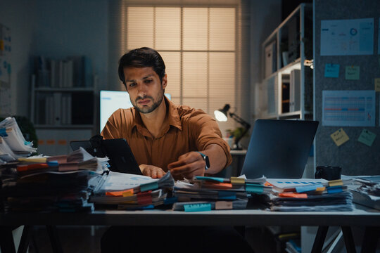 Overworked young Latin businessman working late on document in large stack of paperwork files at office night. Tax audit deadline, accounting stress, corporate burnout, urgent business concept.