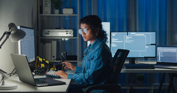 Young African American female programmer coding AI software on multiple screens in night office. Network security, cybersecurity, machine learning, working late, overtime, robotics automation concept.