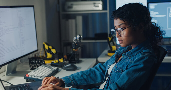 Young African American female programmer coding AI software on multiple screens in night office. Network security, cybersecurity, machine learning, working late, overtime, robotics automation concept.