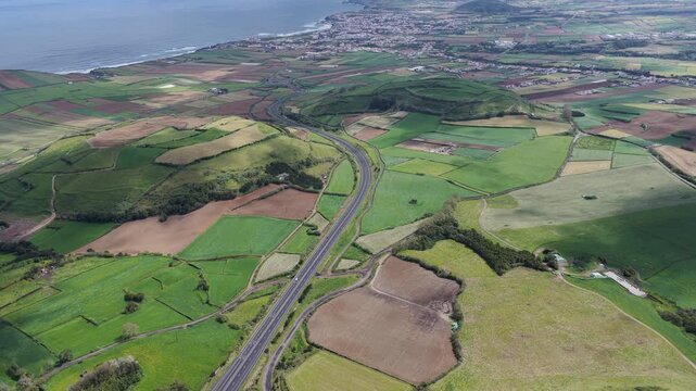 Aerial view of a highway winding through green agricultural fields and rolling hills near the coastline in Rabo de Peixe, Acores, Portugal.
