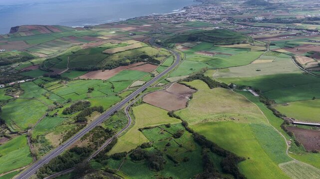 Aerial view of a highway winding through lush green agricultural fields and rolling hills near the coast in Rabo de Peixe, Acores, Portugal.