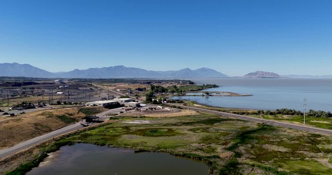 Lindon Marina at Utah Lake, capturing clear blue skies, passing commuter train, calm shoreline, green marshland, and dramatic distant mountains on a sunny day - forward aerial approach
