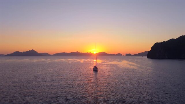 Lone sailboat sits beneath the setting sun while a bright reflection stretches across calm tropical water