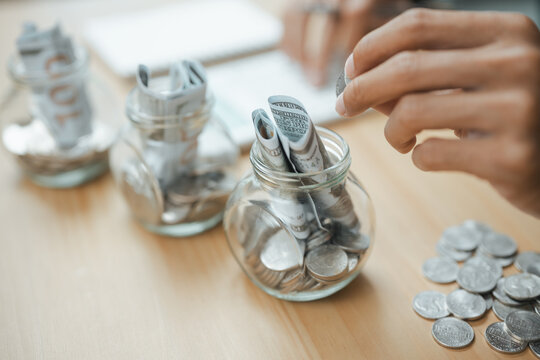 Hand placing coins into savings jars with cash, representing savings goals, budgeting milestones, emergency funds, and personal financial planning for long-term stability and future success.