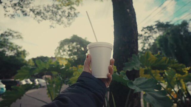 Hand holds a white disposable cup with a straw. Trees and greenery surround the scene. Tree trunk and street background provide depth. The moment captures a relaxed outdoor break