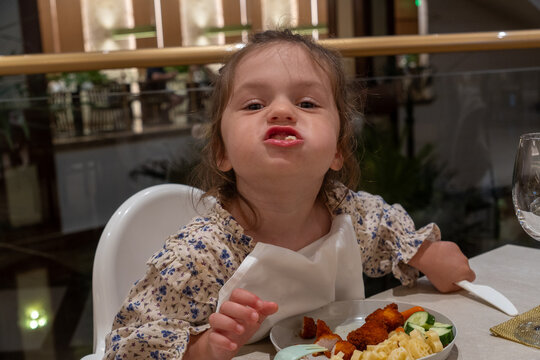 A funny and cheeky little girl sits in a white high chair at a restaurant table, making a silly grimace with her mouth full of food. She is chewing her dinner and looking directly at the camera while
