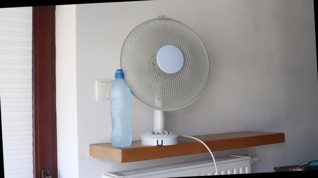 A rotating fan with a frozen plastic water bottle positioned in front of it, illustrating a common home cooling method for hot summer days.