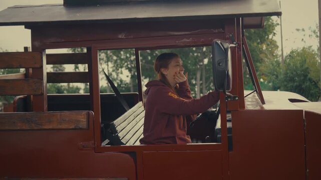 Person in red hoodie talks on phone inside wooden cart. Close-up shows phone in hand, focused communication. Wooden trolley interior has slats and natural light. Green trees visible through window