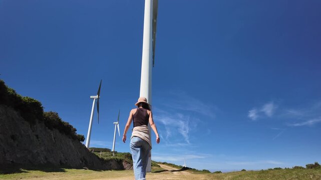 Woman walking below a tall wind turbine on a sunny mountain path