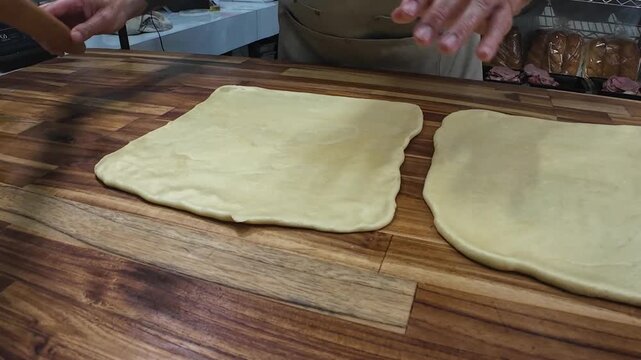 skilled artisan demonstrates the precise technique of rolling dough on wooden table in a commercial bakery setting.