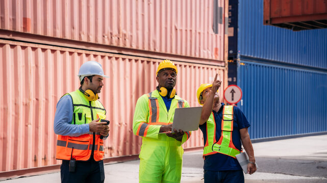 Logistics workers in safety vests and helmets collaborating at a shipping cargo container yard, with a male worker writing on a clipboard during a discussion with his foreman holding a walkie-talkie