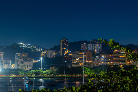 View of Flamengo skyline at night with illuminated high-rise buildings, boats anchored in the bay, and trees in the foreground, Rio de Janeiro, Rio de Janeiro, Brazil.