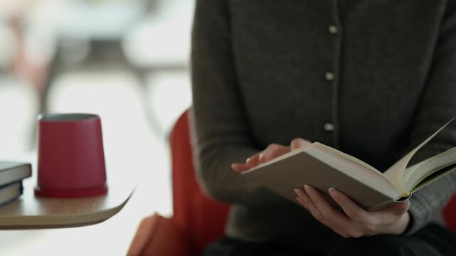 A close-up of a person's hands holding an open book with a red cup and warm blurred background in a cozy reading environment.