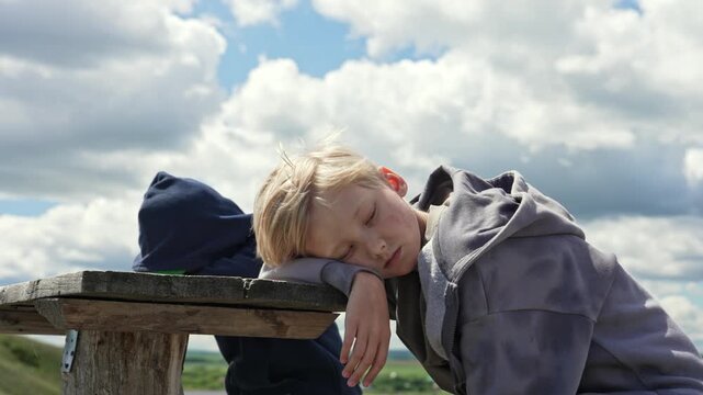 Exhausted young blond boy wearing a grey hoodie sleeping peacefully outdoors on a rustic wooden picnic table after a long day of playing, with a beautiful cloudy sky in the background