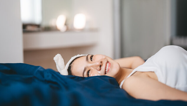 Beautiful smiling young asian woman, relax lying in bed with eye sheet mask enjoy skincare wellness routine for positive self care